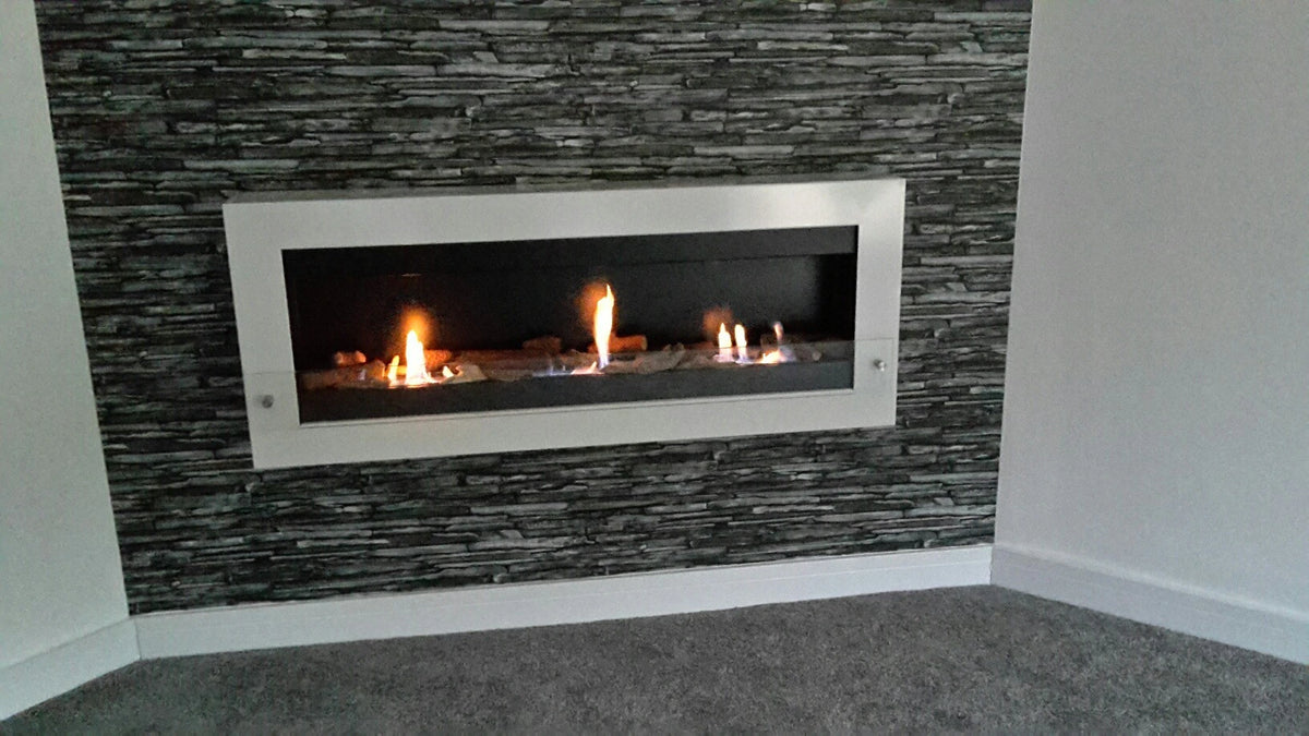 XL white-framed wall-mounted bioethanol fireplace with lit flames, styled against a dark stacked-stone accent wall with grey carpet flooring