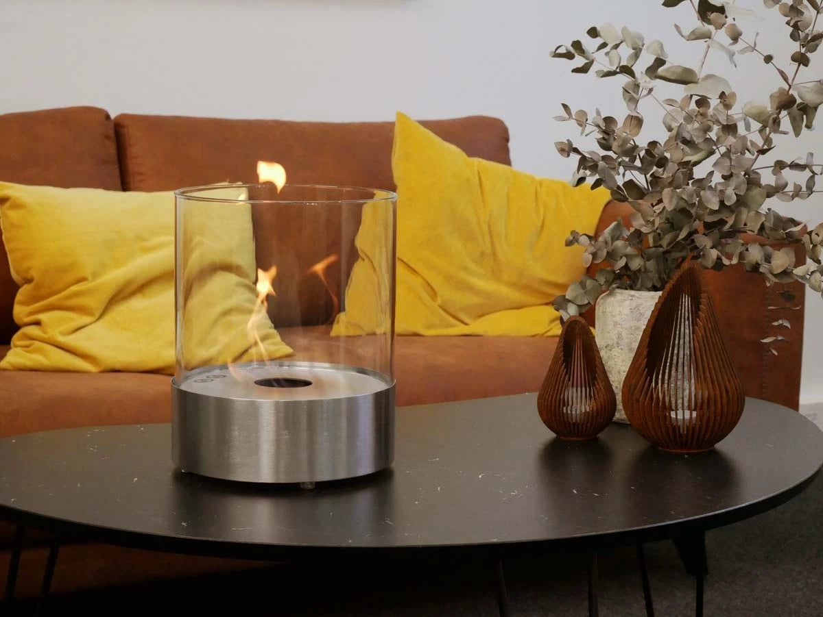 Southwark round tabletop bioethanol fireplace in steel, lit flame, on a coffee table with sofa and dried eucalyptus decor