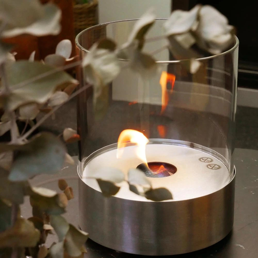 Close-up of the Southwark round tabletop bioethanol fireplace in steel, lit flame, with eucalyptus branches