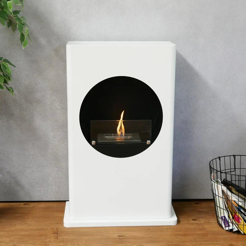 White freestanding bioethanol fireplace with round cutout in a living room with a plant and magazine basket, lit flame