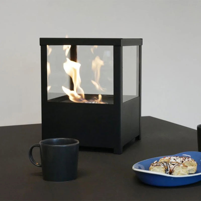 Square black tabletop bioethanol fireplace lit on a dark table with a coffee mug and pastry