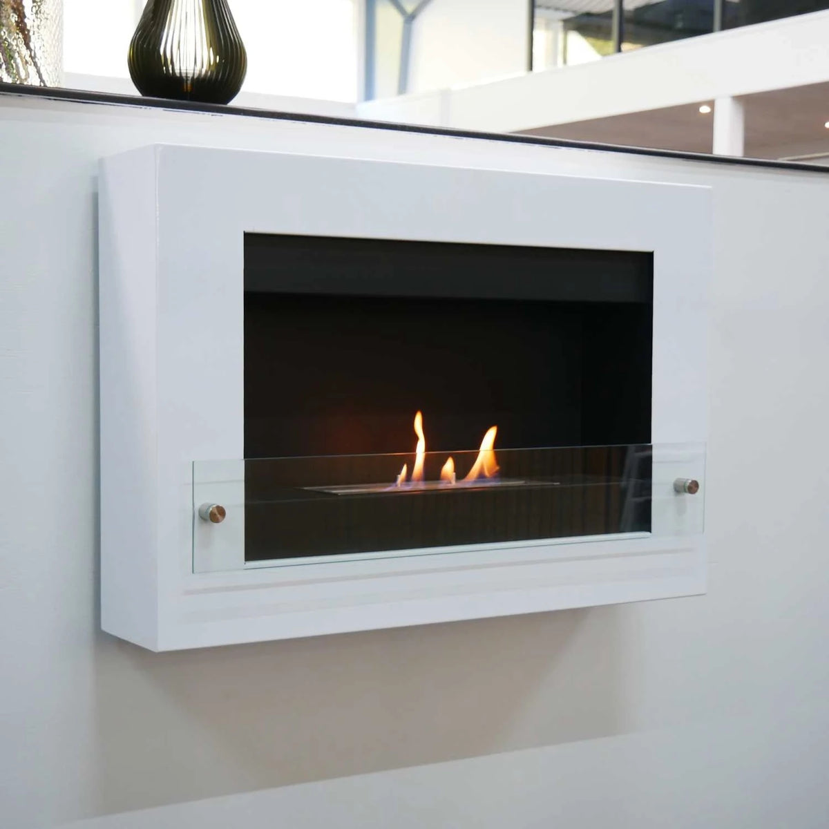 White wall-mounted bioethanol fireplace lit in a modern room with a vase on a shelf above it