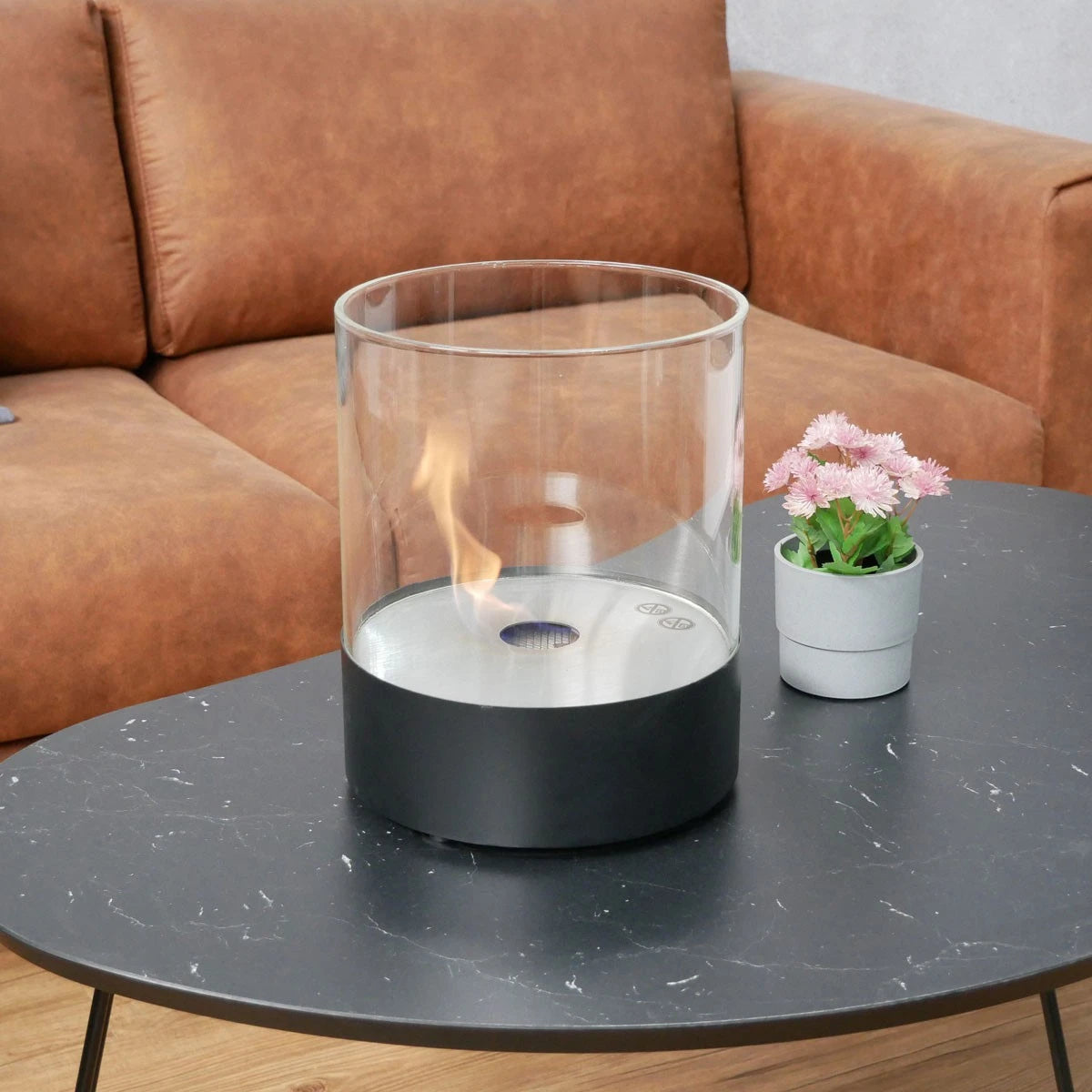 Southwark black round tabletop bioethanol fireplace on a marble coffee table with a tan sofa and pink flowers, lit flame