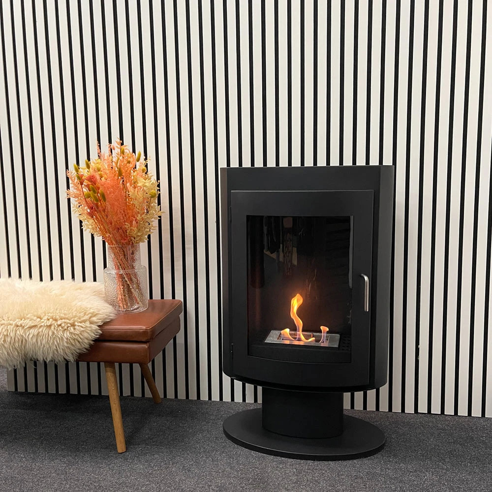 Trenton bioethanol stove fireplace against a striped wall, lit flame, with a leather stool and dried flowers