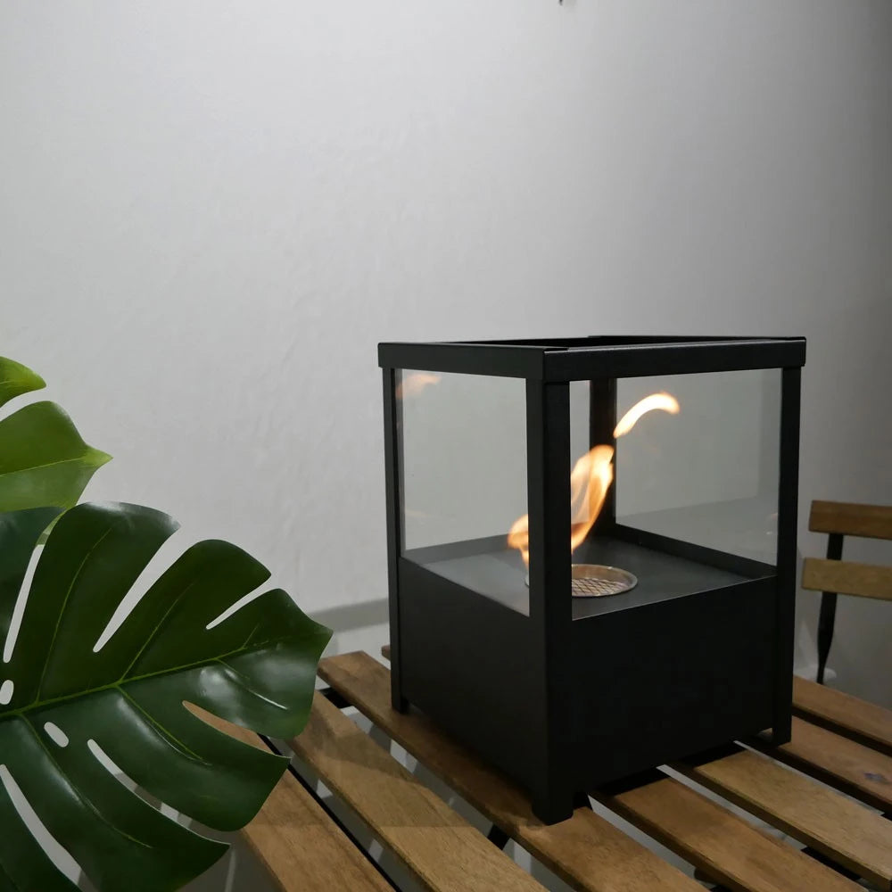 Square black tabletop bioethanol fireplace lit on an outdoor wooden table beside a plant