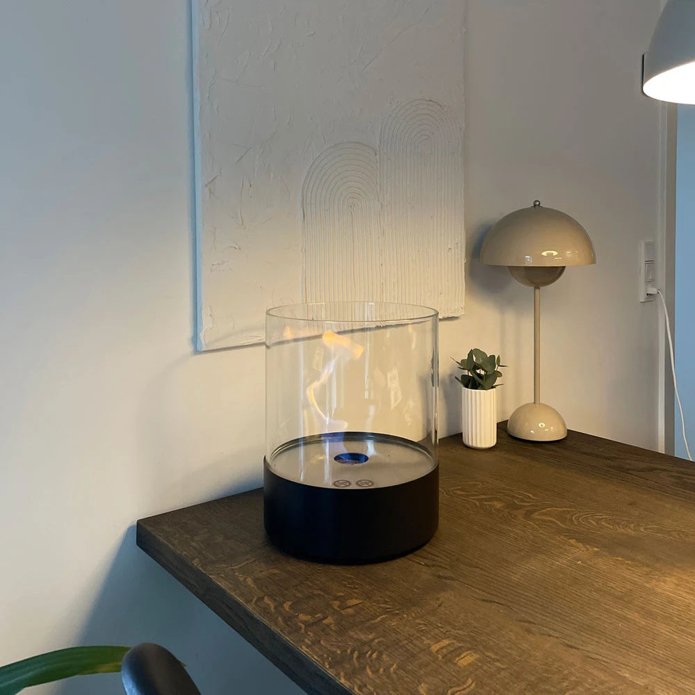 Southwark black round tabletop bioethanol fireplace on a wooden dining table with a lamp and plant, lit flame