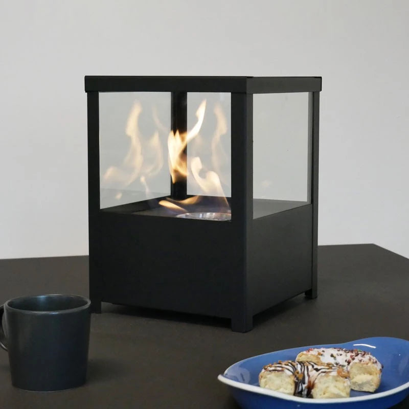 Square black tabletop bioethanol fireplace lit on a dark table with a coffee mug and pastry, wider view