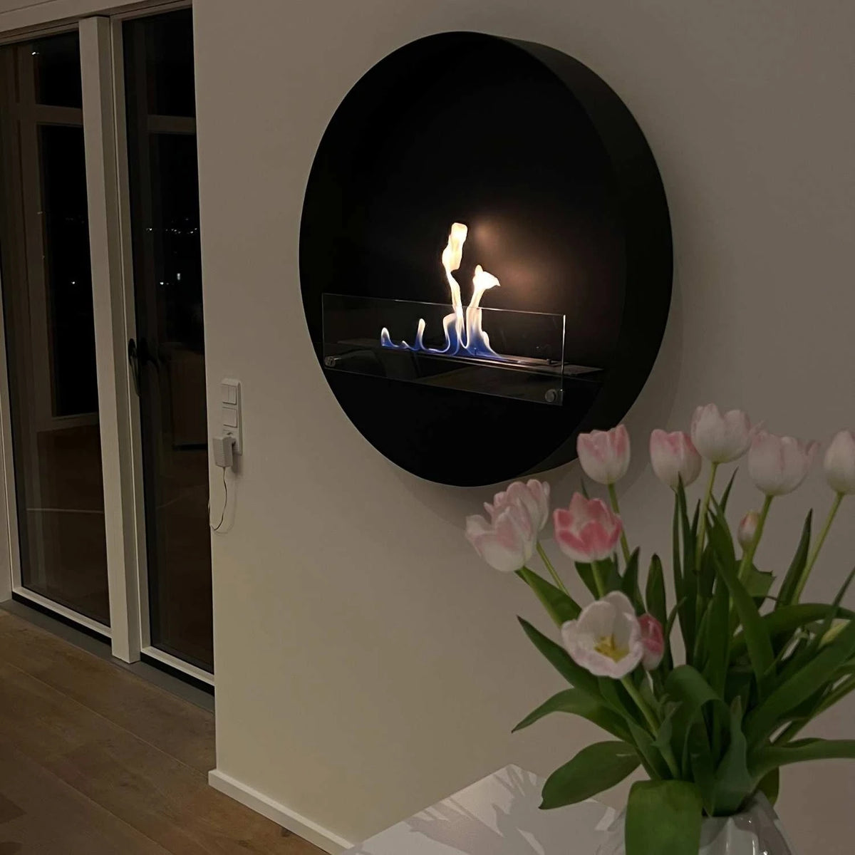 Delaware XL black round bio fireplace mounted in a hallway at night, lit with a realistic flame, tulips in the foreground